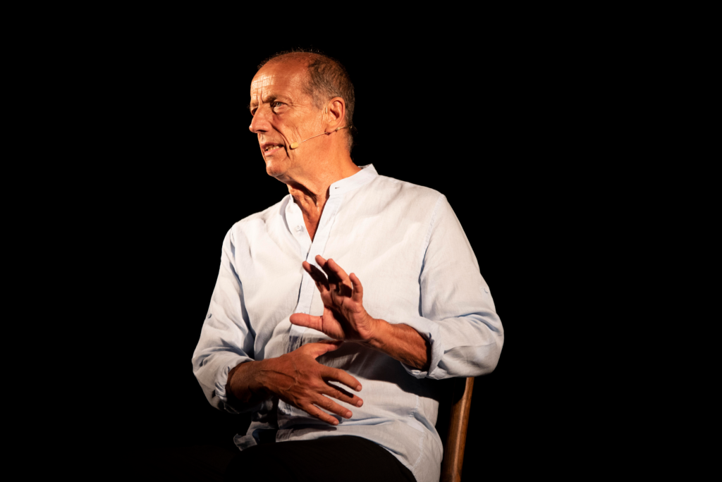 Un uomo da solo seduto su una sedia parla attraverso un microfono ad archetto. Ha una camicia bianca con un collo alla coreana. L'uomo ha circa 70 anni ed ha i capelli rasati ed è un po' stempiato.